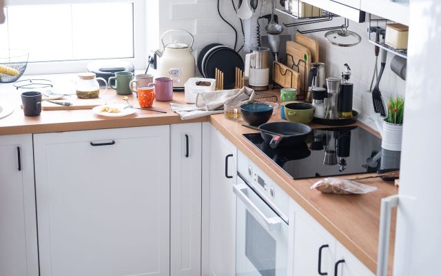 dirty kitchen with mugs, pans, trash, food laying on counters.