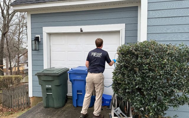Jeffcoat tech spraying or painting a white garage door on a light blue house, with trash bins nearby.