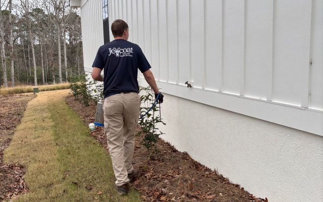 Jeffcoat tech in a “Jeffcoat Landscape Services” shirt walking beside a white building with shrubs and pine straw mulch.