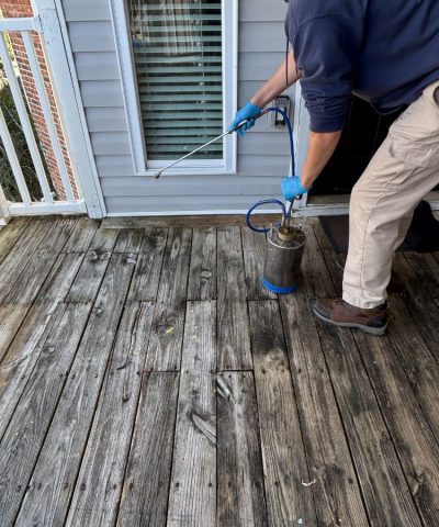 Jeffcoat tech applying liquid treatment to a weathered wooden deck using a metal sprayer, wearing gloves and a navy hoodie.
