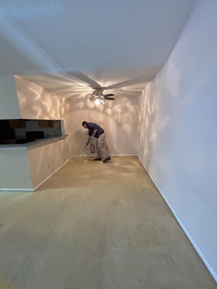 residential-pest-control-services-columbia-sc Jeffcoat tech using a handheld sprayer on beige carpet in a mostly empty room with patterned ceiling shadows and a kitchen pass-through.