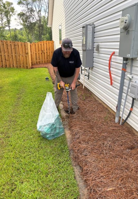 termite-treatment-bait-traps-lexington-sc-pest-control Jeffcoat tech drill hole into ground for termite bait station next a house with white siding and utility boxes, near mulched soil.