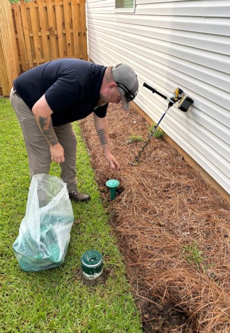 termite-treatment-bait-traps-newberry-sc-pest-control Jeffcoat tech putting termite bait station into ground for next a house with white siding.