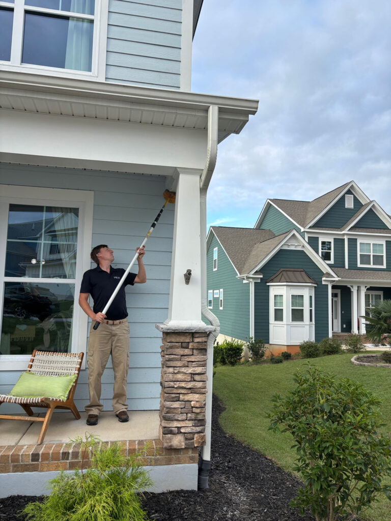 Jeffcoat tech porch cleaning roof eaves with long brush; light blue house, stone column, green chair nearby.